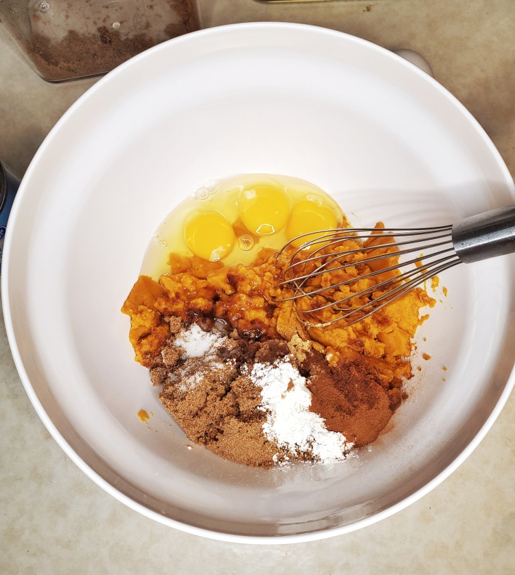 Picture of an bowl with pumpkin puree, spices, sugar, 3 eggs inside. A whisk is seen resting in the bowl.